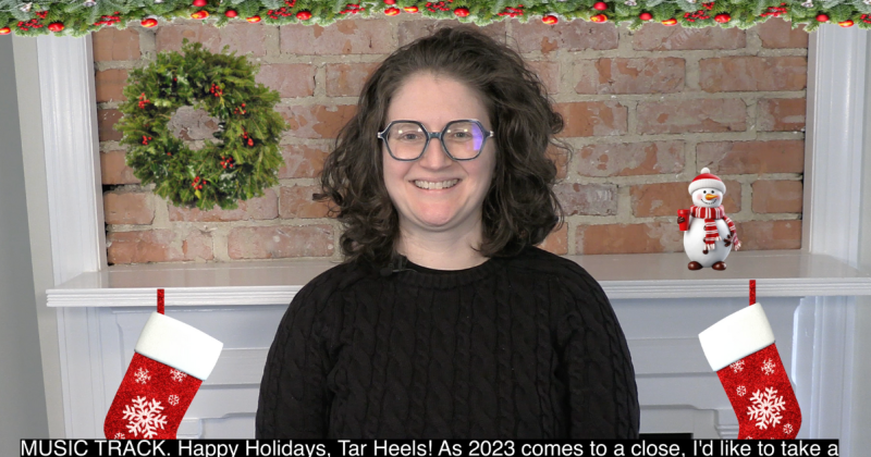 Elizabeth Hall smiles during her holiday video address. Behind her is a fireplace with stockings, a holiday wreath and a snowman.